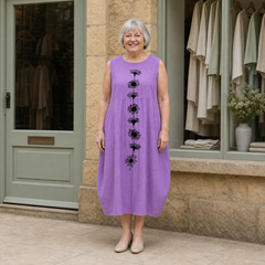 Margaret models the violet Emma midi dress with a black daisy-stem design in a front full-body view outside a pale-stone Cotswold boutique.
