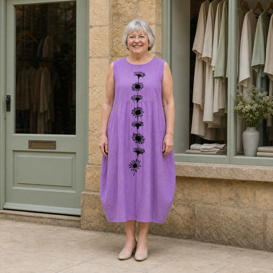Margaret models the violet Emma midi dress with a black daisy-stem design in a front full-body view outside a pale-stone Cotswold boutique.