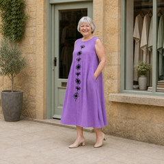Margaret models the violet Emma midi dress with a black daisy-stem design in a slight three-quarter full-body view outside a pale-stone Cotswold boutique.