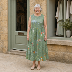 Margaret models the sage Hope woodland midi dress with illustrated animals and mushrooms in a front full-body view outside a pale-stone Cotswold boutique.