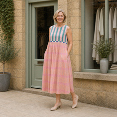 Helen models the pink and teal striped Lily pastel midi dress in a slight three-quarter full-body view outside a pale-stone Cotswold boutique.